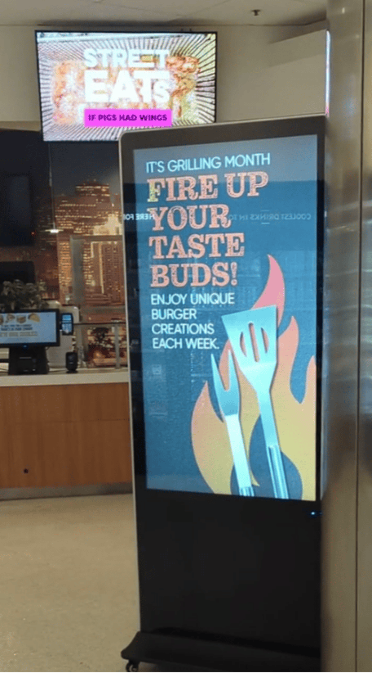 Freestanding digital poster kiosk showing a Fire Up Your Taste Buds burger promotion in a food court