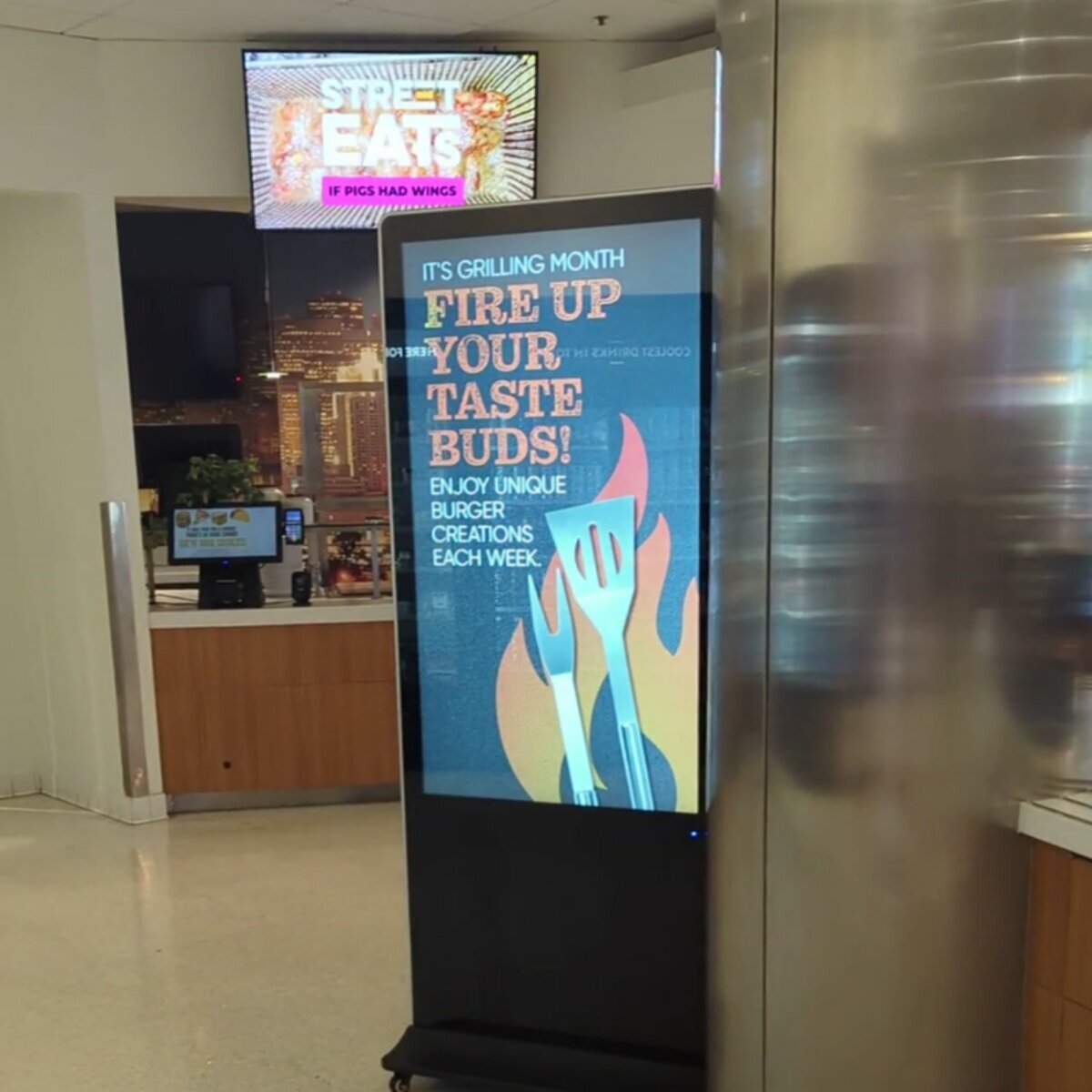 Freestanding digital poster kiosk showing a Fire Up Your Taste Buds burger promotion in a food court